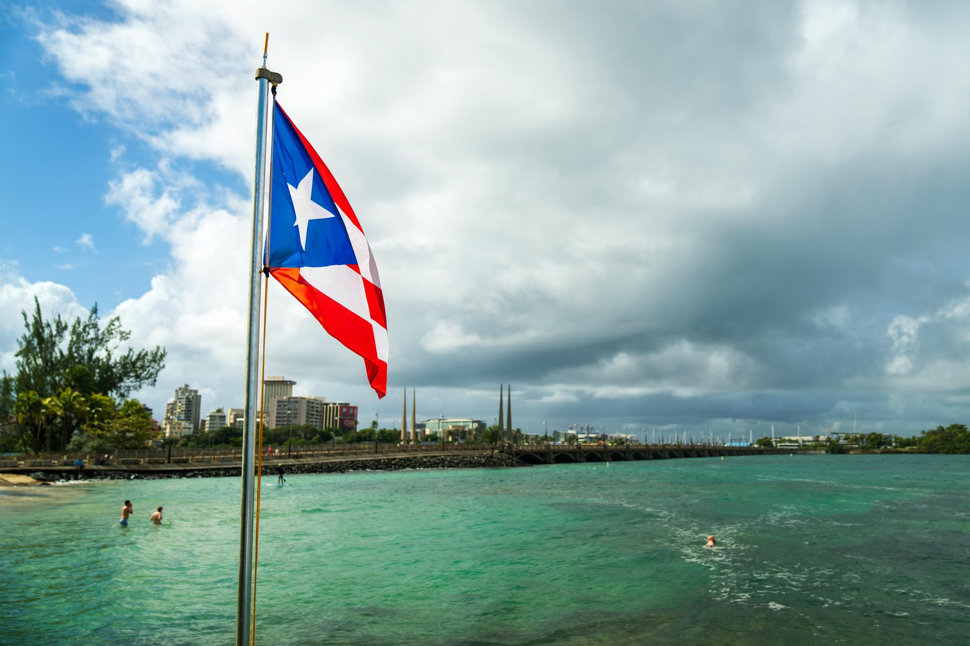 The Puerto Rican flag flying over Condado Lagoon in San Juan, with swimmers in turquoise water and the city skyline behind